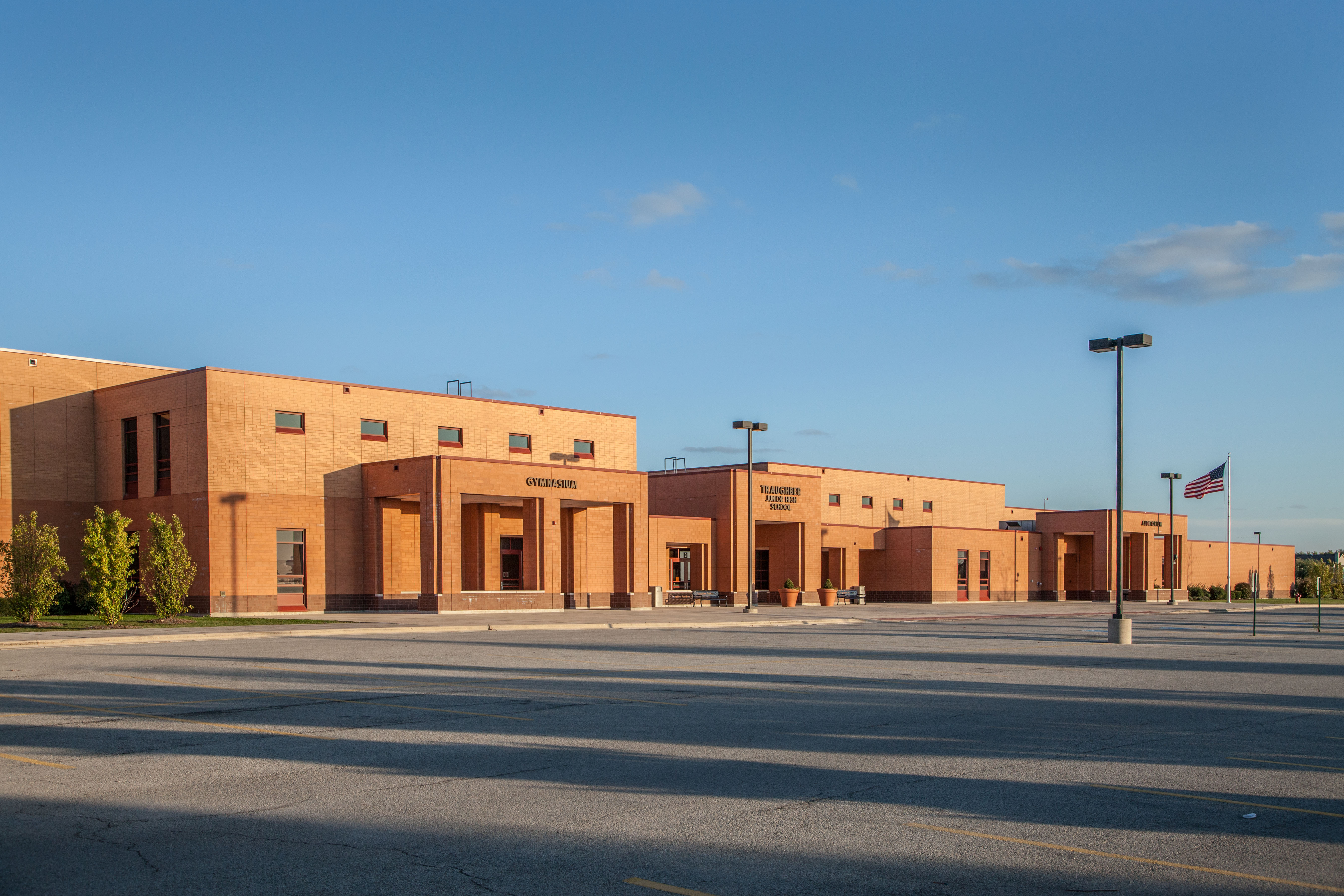 Front of Traughber Junior High showing the parking lot, American Flag pole on right side, entrances for the Gym (left), main entrance (center), auditorium (right), and a blue sky in the background. 